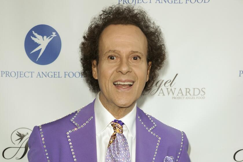 A man with short curly hair in a purple suit with a bedazzled trim and a matching tie smiling with his mouth open