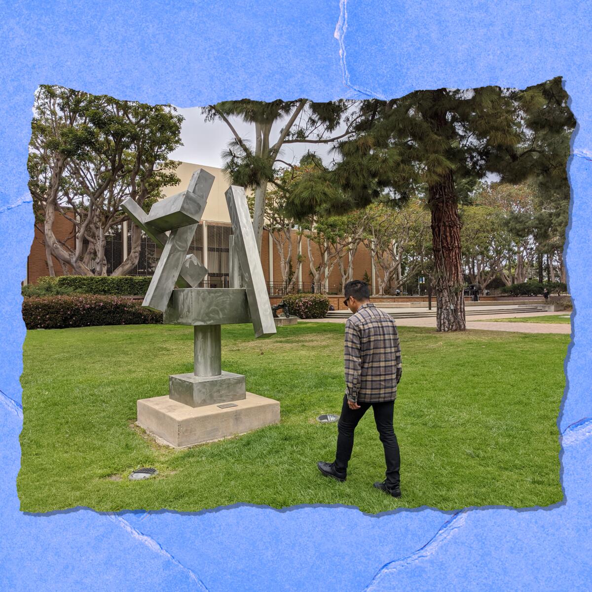 A person stands in front of a sculpture on a green lawn. Trees, shrubs and a brick building are in the background.