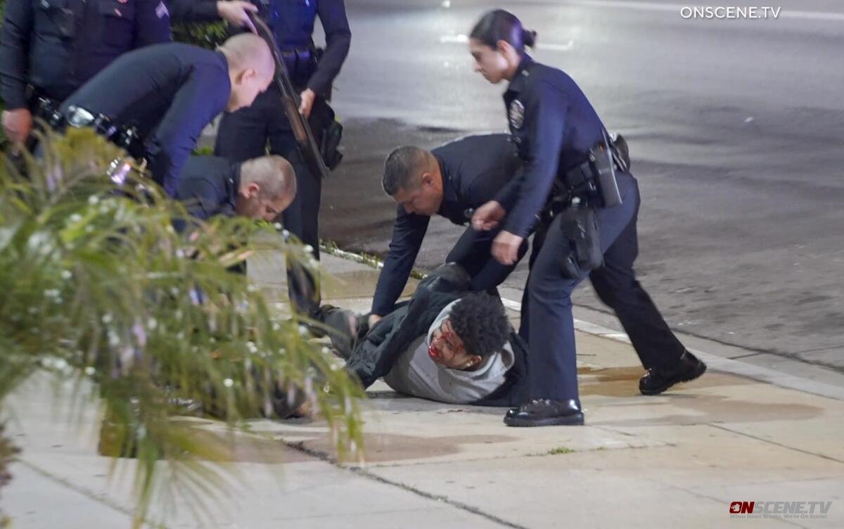 Police officers surround and handcuff a person lying on the sidewalk with blood on his face.