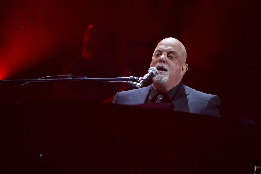 A bald man with a gray beard wearing a suit while singing into a microphone and playing piano