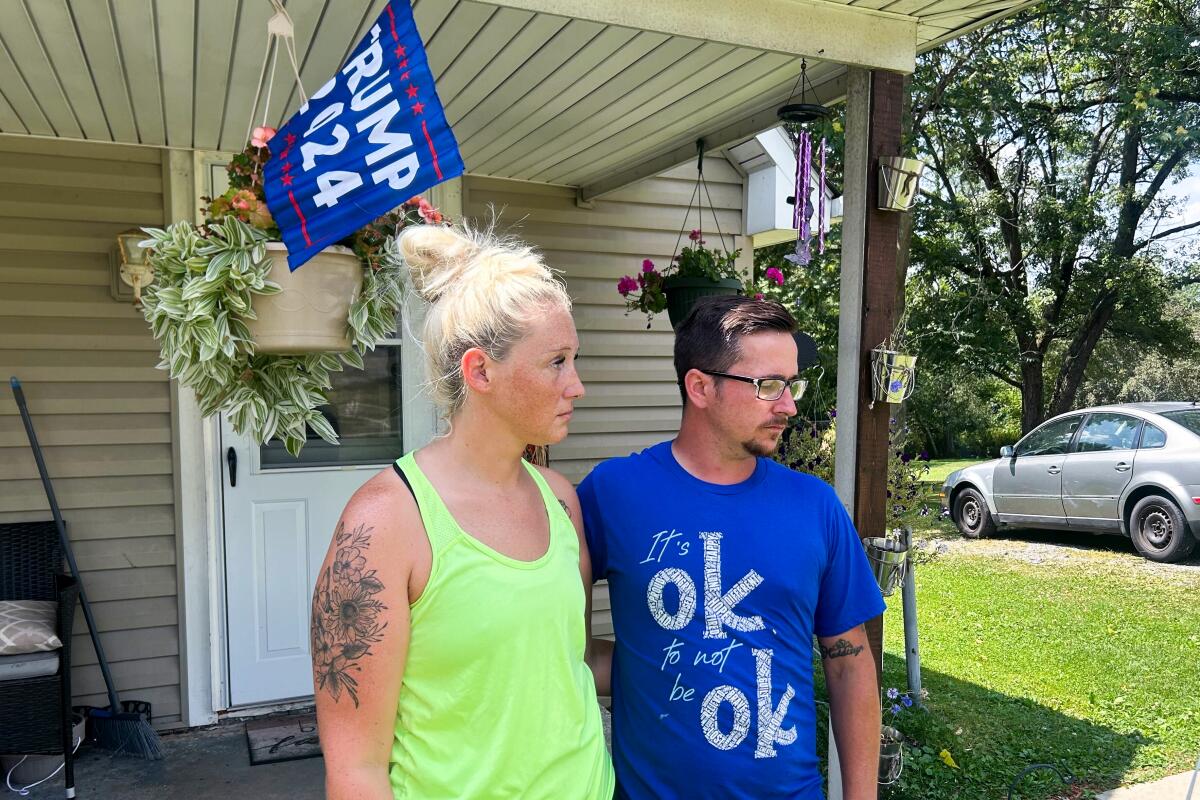 Jessica Lynch and her fiancé Robert Runyan stand in front of a house with a Trump 2024 flag hanging over the porch.