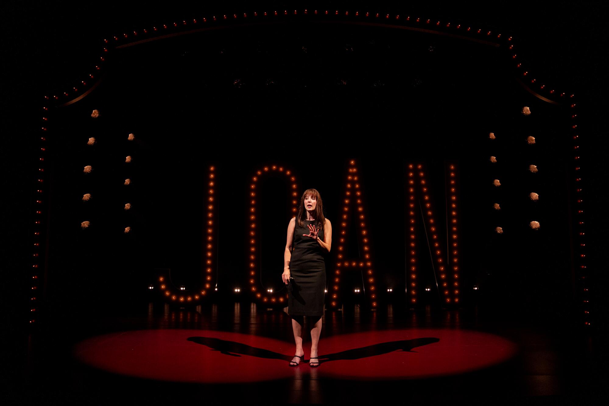 A woman stands onstage holding a microphone, in front of lights that spell out JOAN.