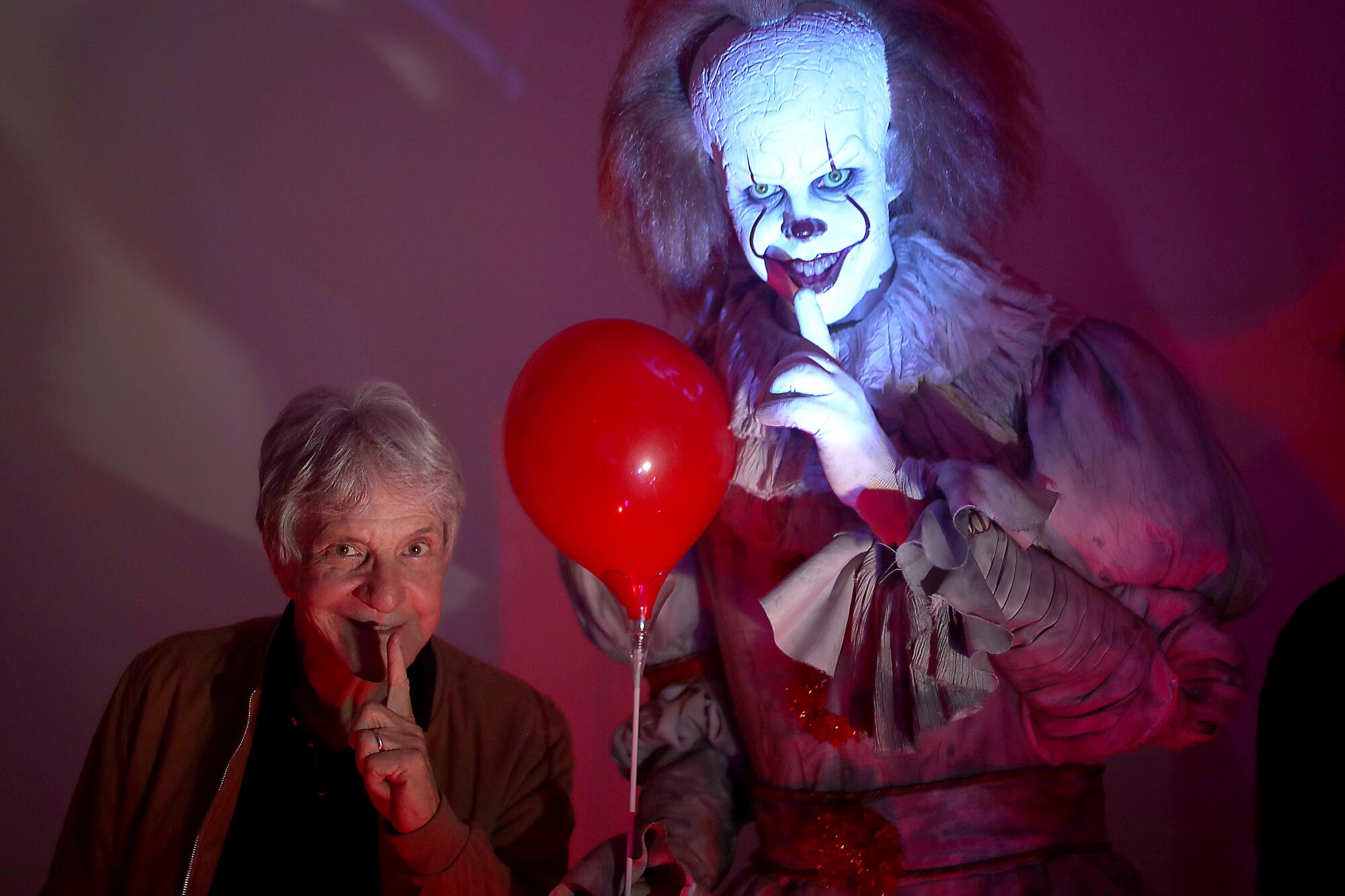 A man at left holds a finger to his lips. That's duplicated by a person to the right of a balloon with scary clown makeup.