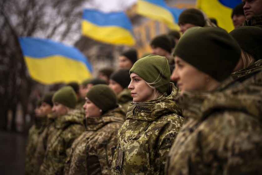 A group of female soldiers in camouflage stand near others and blue and yellow flags