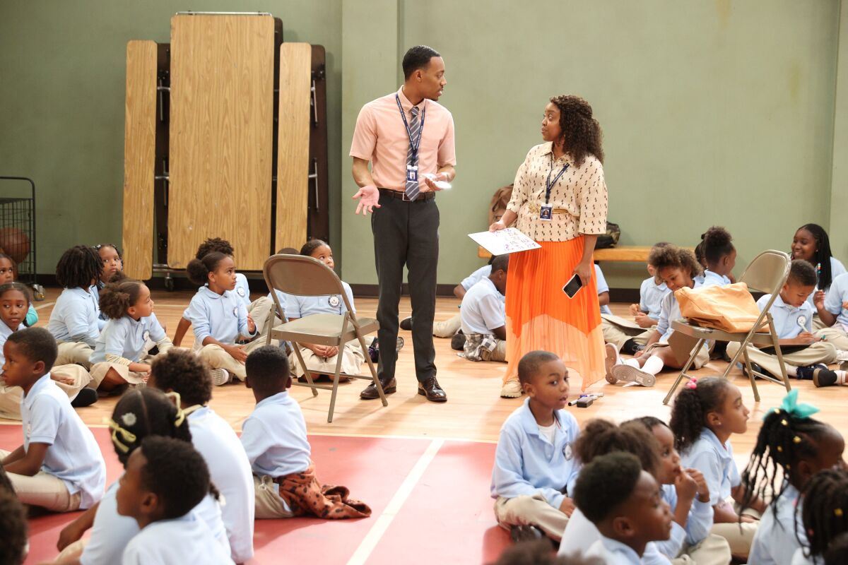 Two teachers standing in a room surrounded by children seated on the floor