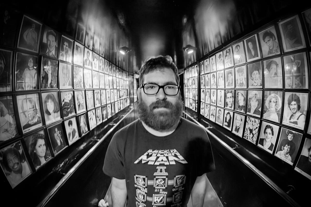 A man with a thick beard and glasses stands in a hallway lined with framed portraits.