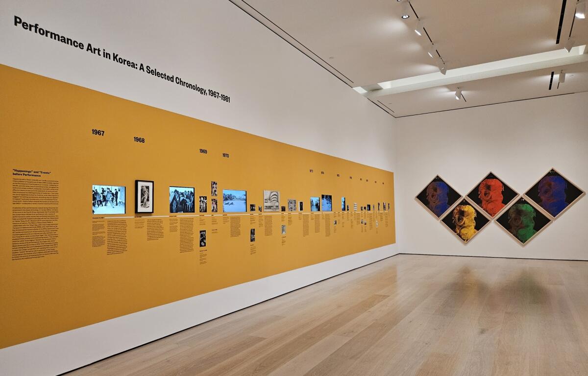 A timeline of still and video images is  displayed on a yellow museum wall. Five silkscreen images hang beside it.