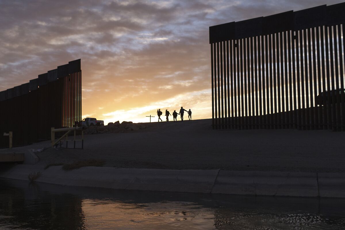 A small group of people are silhouetted as they walk through a large gap in the border wall