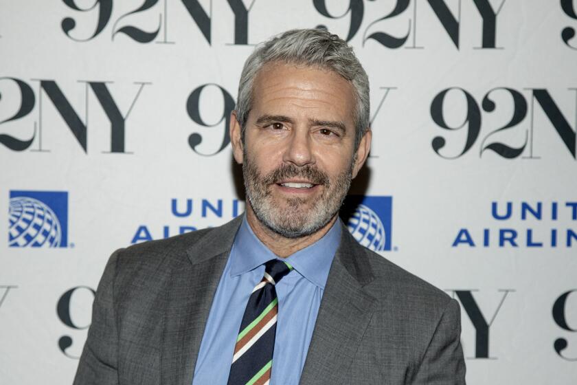 Andy Cohen in grey suit and multicolor tie, posing in front of a 92NY backdrop