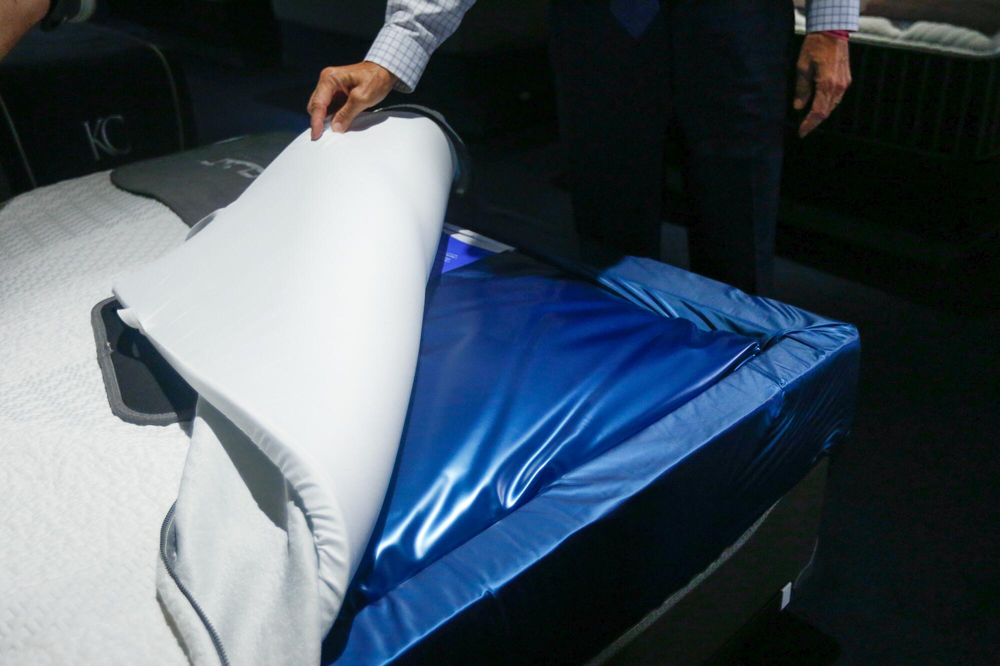 A hand lifts part of a mattress to reveal a waterbed.