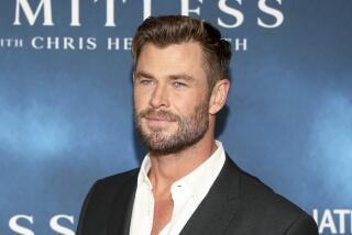 Chris Hemsworth in a black blazer and white suit shirt posing in front of a blue backdrop