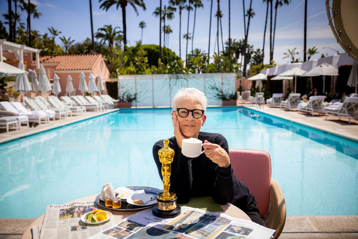 A woman sits poolside holding a cup of coffee, an Oscar on the table next to her.