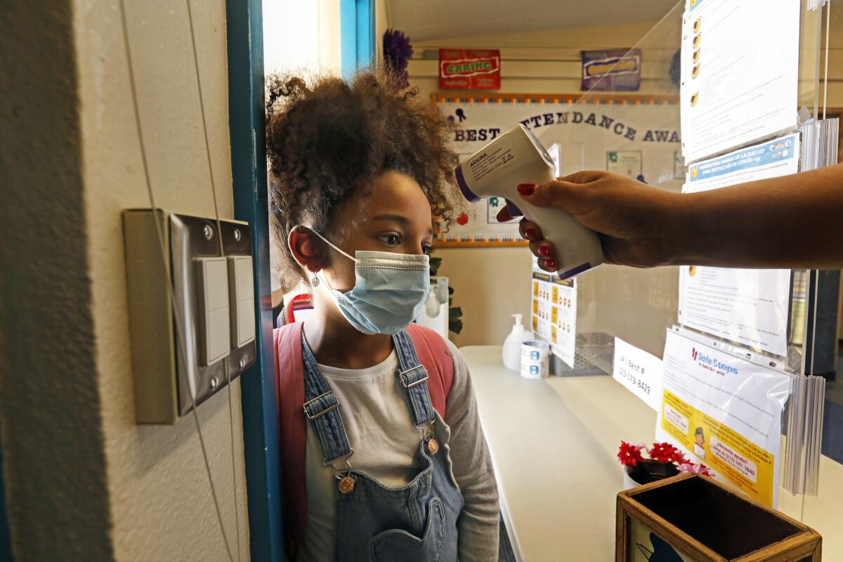 A girl in a mask and a backpack stands as she's scanned with a forehead thermometer.