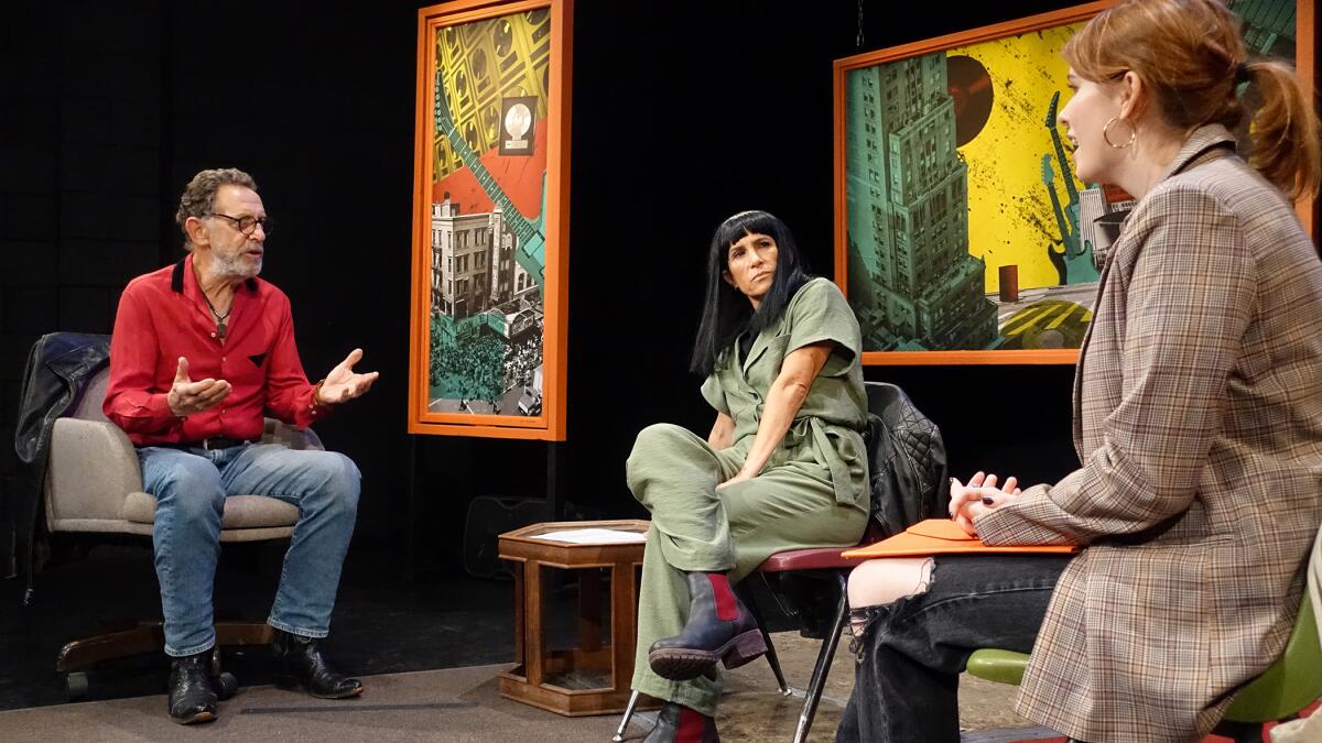 A man and two women talk as they sit in chairs on a stage. In the background are two framed artworks and a black backdrop.