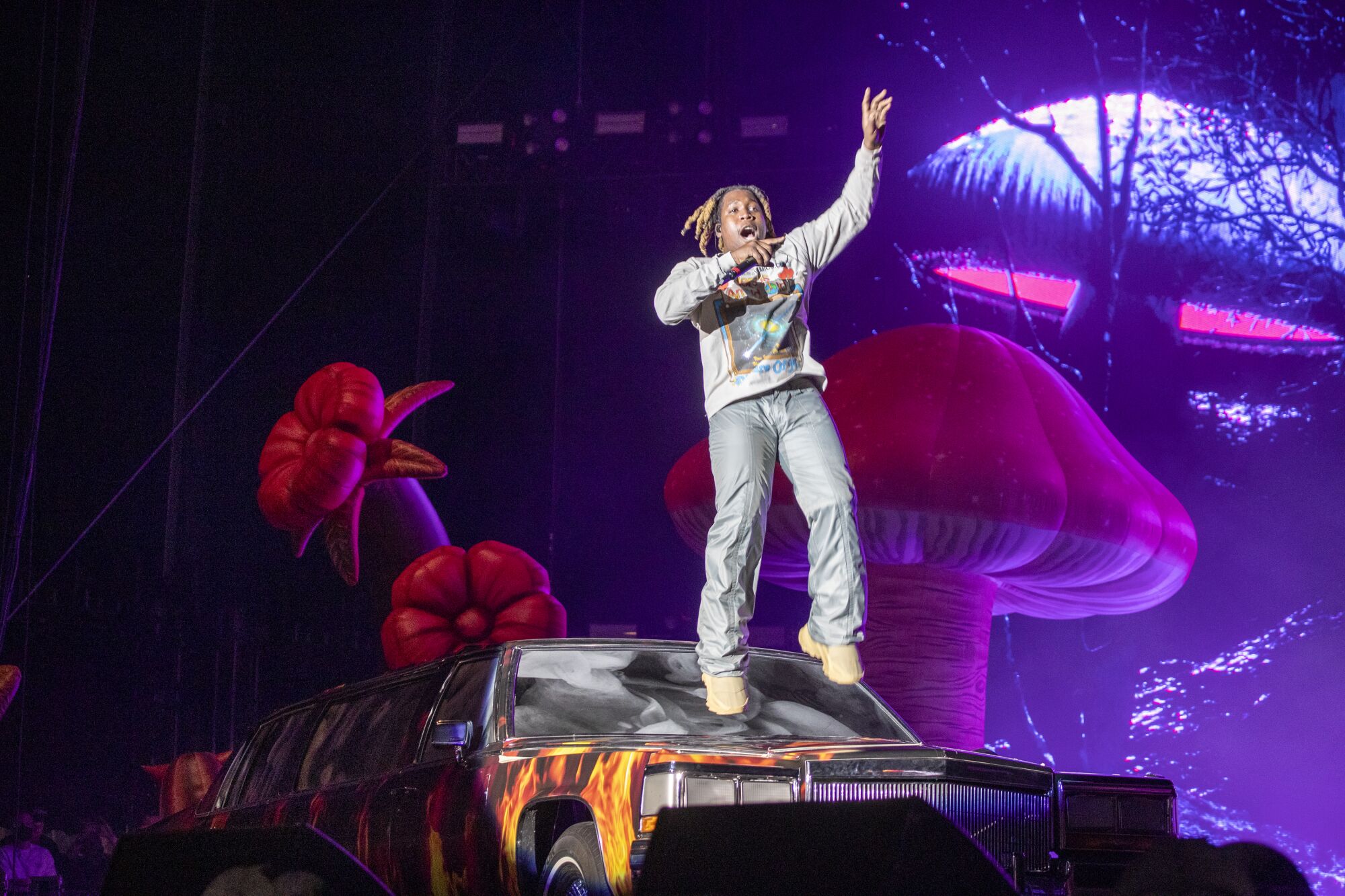 A man jumps on a car onstage with a giant mushroom and flowers in the background