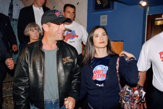 An old image of Bruce Willis in a hat and leather jacket holding hands with Demi Moore in a black shirt