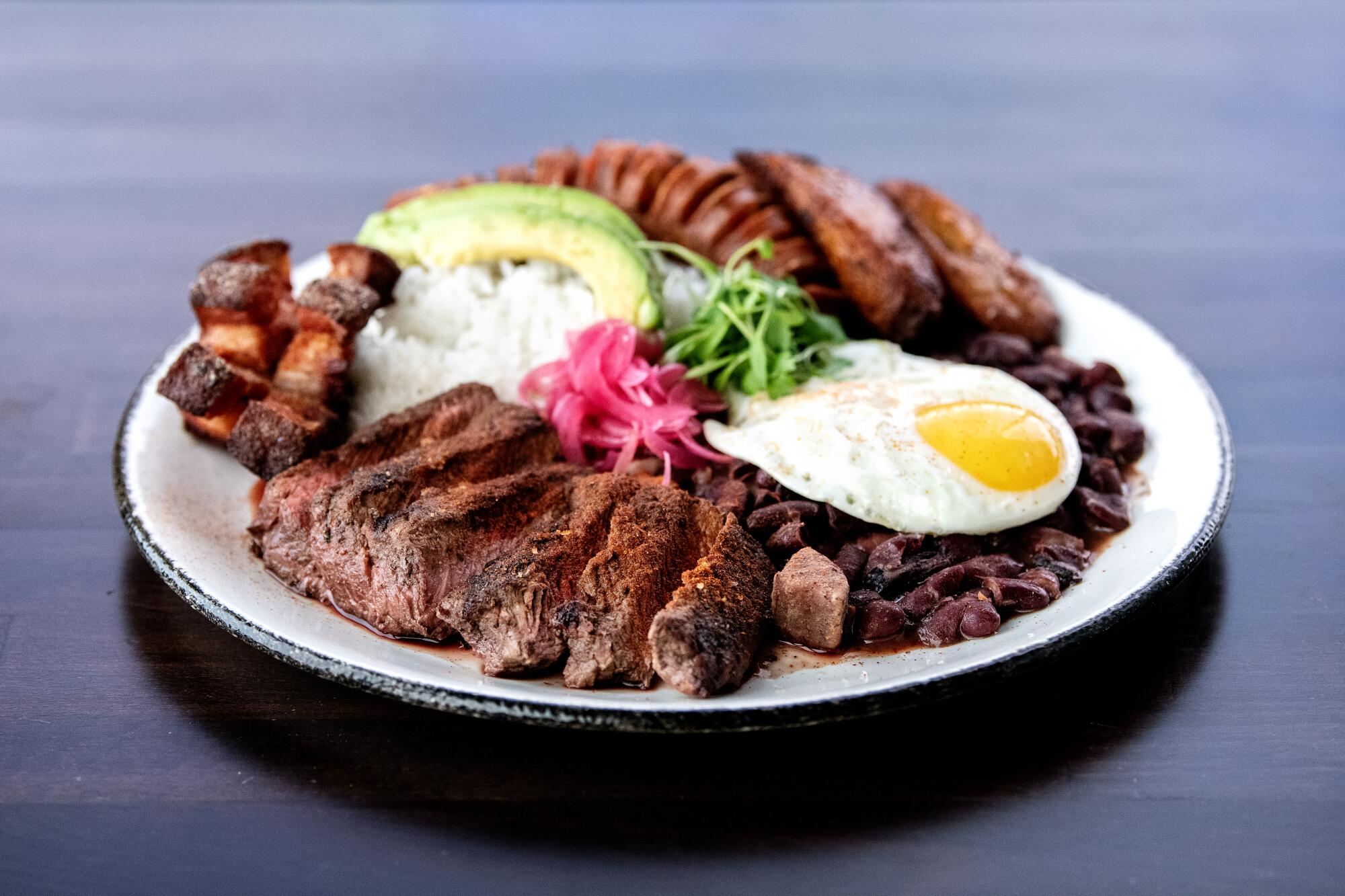 A plate with a variety of meats, plus rice, a slice of avocado and a fried egg.