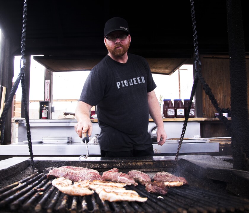 A man wearing a black T-shirt with the word "pioneer" mans an outdoor grill.