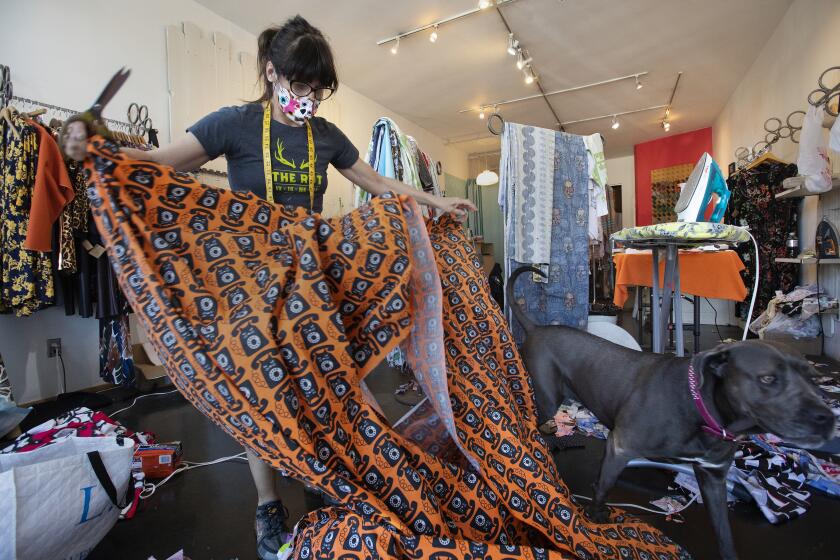 SILVER LAKE, CA-APRIL 22, 2020: Laura Howe, owner and designer at Matrushka Construction, a women's boutique in Silver Lake, works with telephone themed fabric, that is being used to make masks. After running out of scraps from fabric used to make dresses, she is now buying fabric, like this one at the garment district in downtown Los Angeles, specifically to be used for making masks. At right is her dog Baby, an Italian Mastiff. (Mel Melcon/Los Angeles Times)
