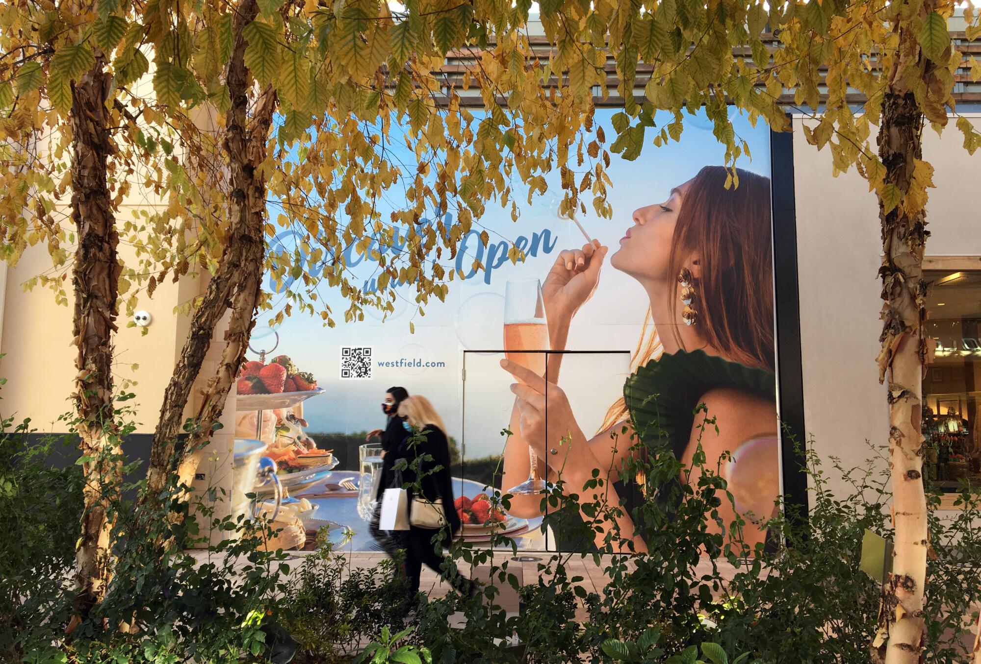Shoppers in masks walk past an advertising poster of a woman drinking from a champagne flute and blowing bubbles.