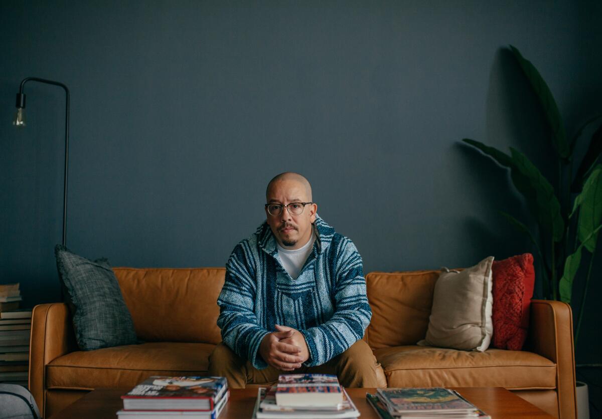 A man sits on a couch, a coffee table laden with books in front of him