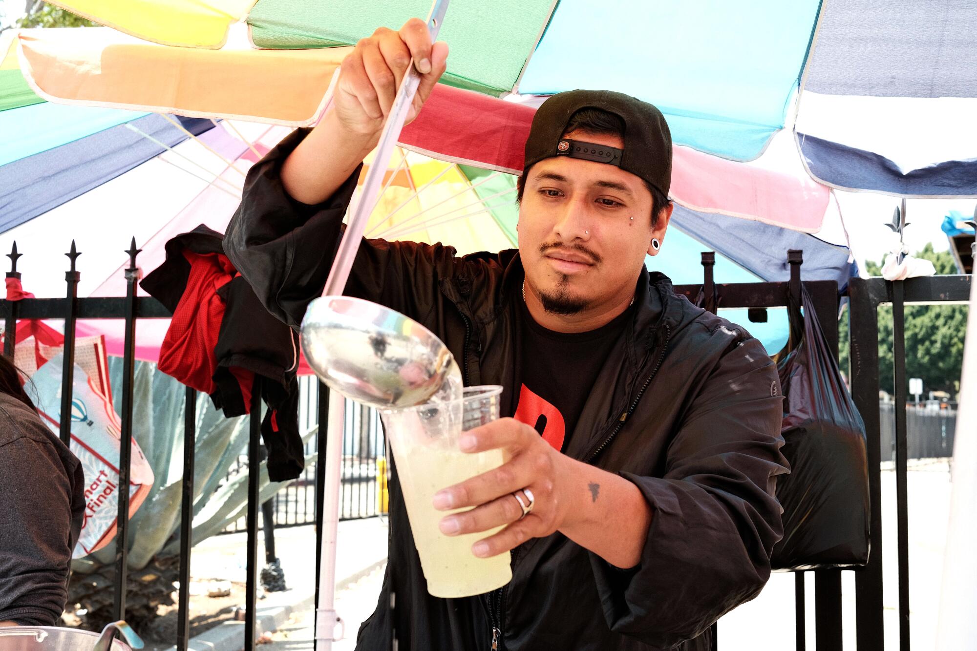 A man in dark clothes ladles a drink into a clear cup under an umbrella