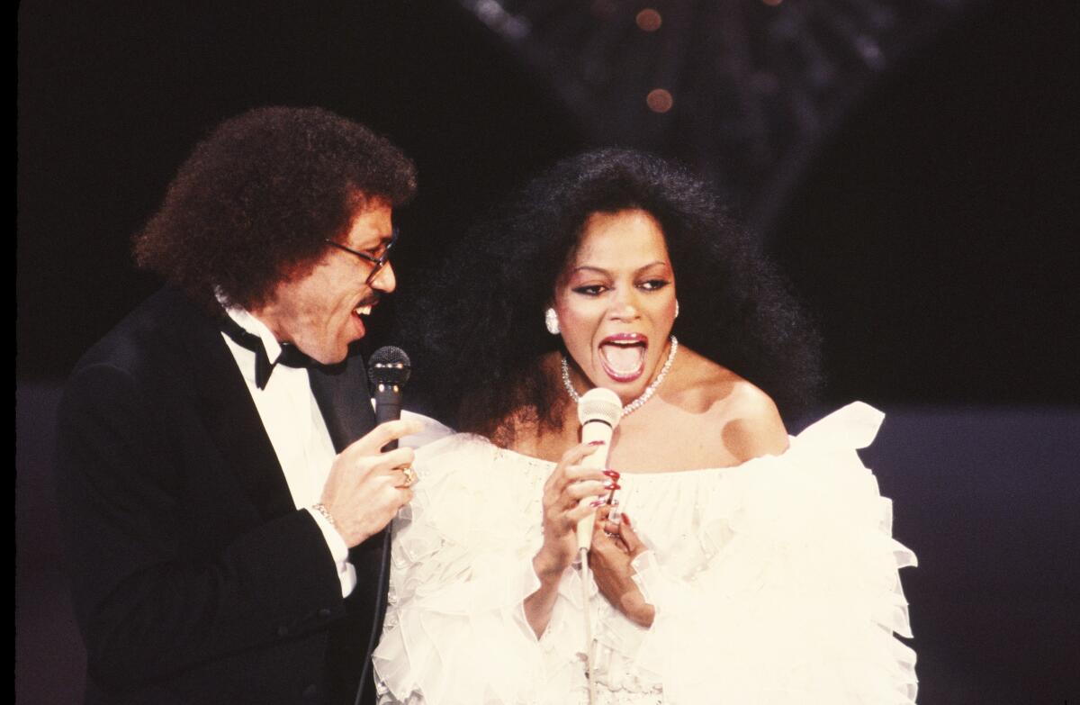 Lionel Ritchie in a tux and Diana Ross in a white gown sing into microphones standing beside each other