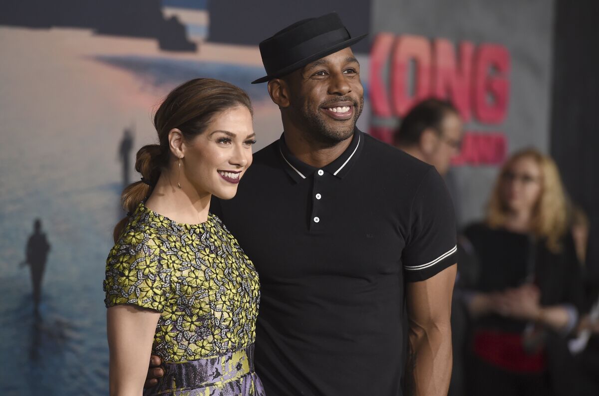 A woman with long brown hair in a yellow dress posing with a man in a black hat and black shirt, both smiling