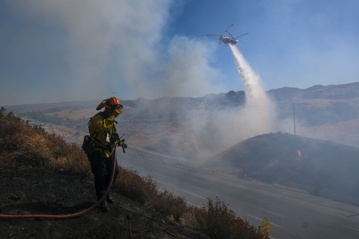 A firefighter sprays a hose on a hillside. In the background, water falls from a helicopter.
