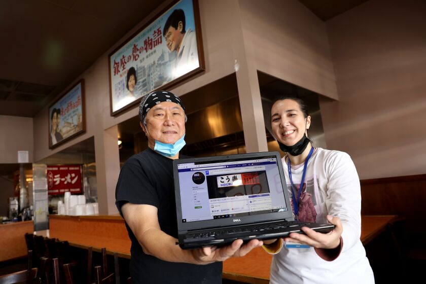 LOS ANGELES, CA -- APRIL 08: Lowering their masks for a photo, Hiroshi Yamauchi, owner, left, with Mariko Lochridge, small business counselor, of Little Tokyo Service Center,showing his restaurant's Facebook page at Kouraku Restaurant in Little Tokyo on Wednesday, April 8, 2020, in Los Angeles, CA. Hiroshi Yamauchi, owner, is learning how to organize and perform take-out food through social media and digital devices for the first time in the history of the restaurant from Mariko Lochridge, small business counselor, of Little Tokyo Service Center. (Gary Coronado / Los Angeles Times)