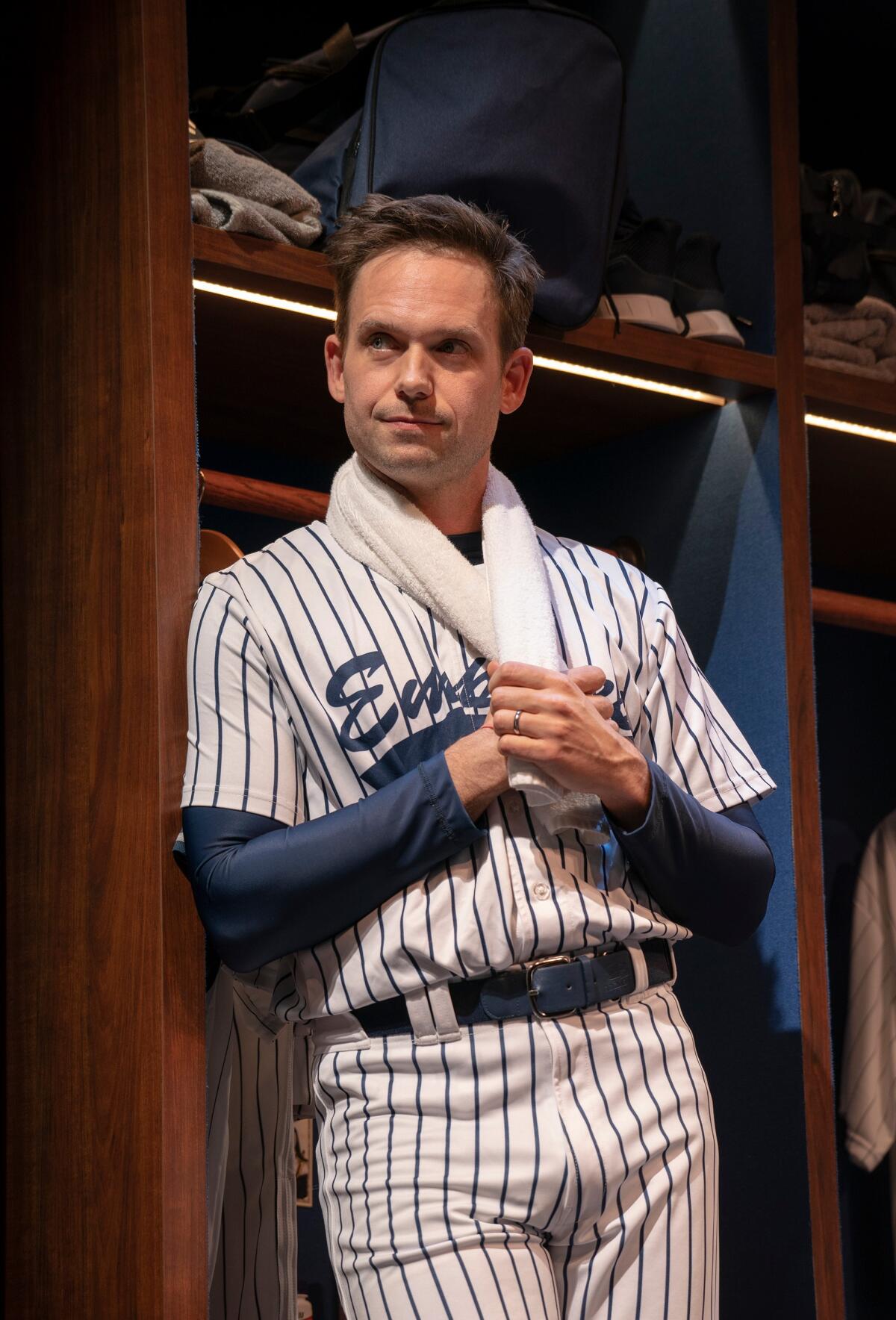 A man in a baseball uniform with a towel around his neck leans against a locker room wall.