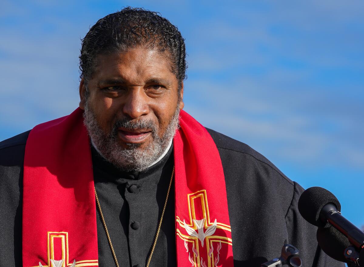 The  Rev. William Barber speaking in front of a microphone while wearing a black robe and red sash