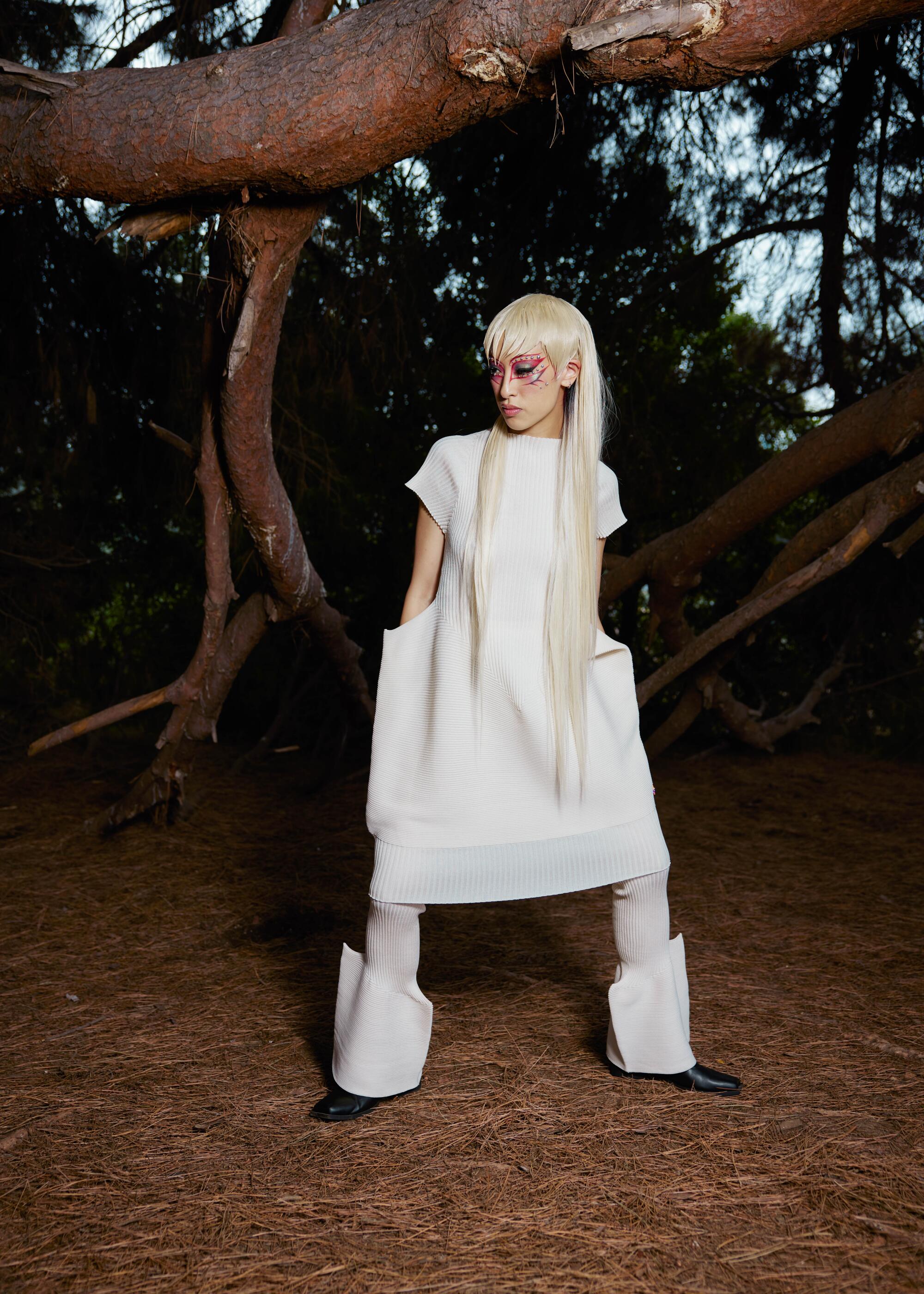 A person with long white-blond hair stands outside in a white Issey Miyake outfit.
