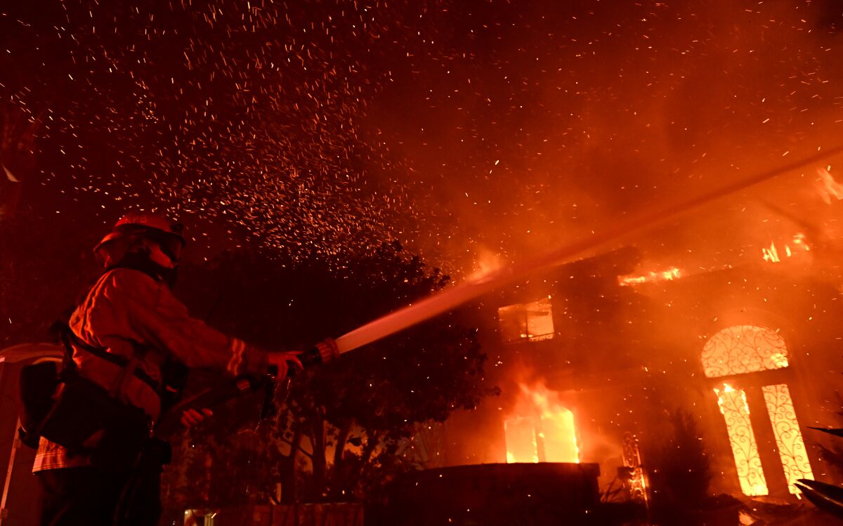 A firefighter sprays water on a burning house at night as sparks fill the air