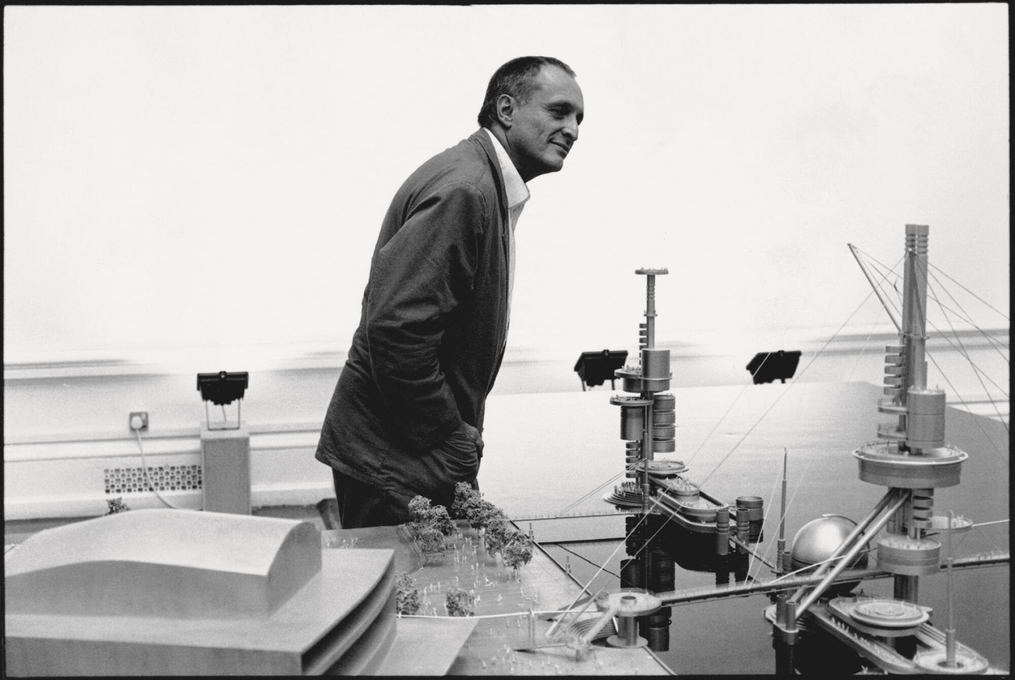 Architect Richard Rogers walks behind a table of architectural models in a black and white photo