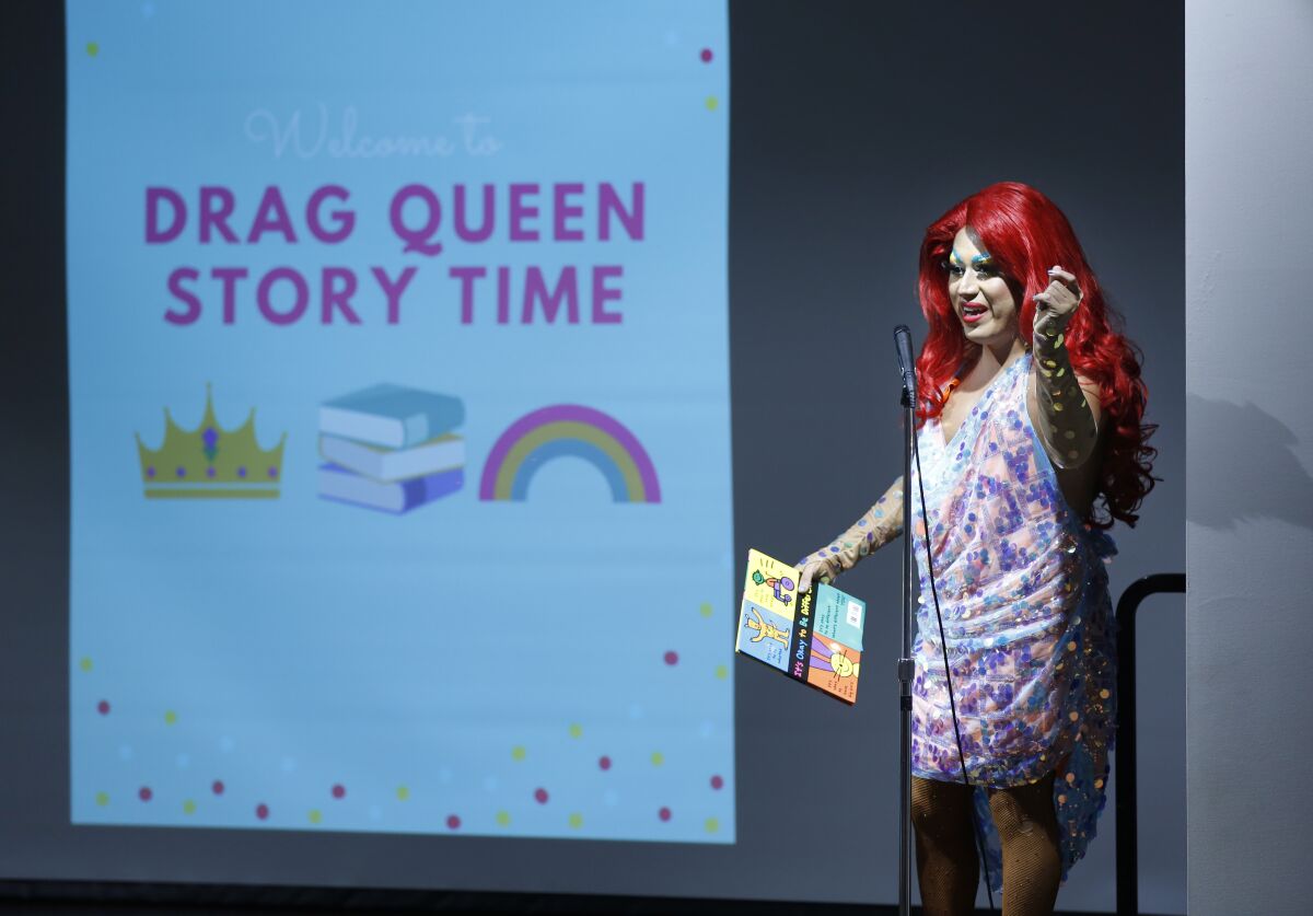 Raquelita, a drag queen, stands next to a sign that says "Drag Queen Story Time" 