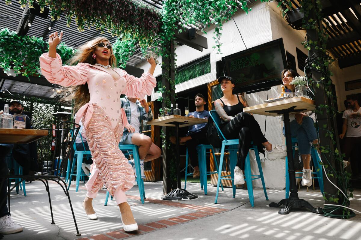 A drag queen dressed in pink strides past people seated at a bar and at tables on a patio.