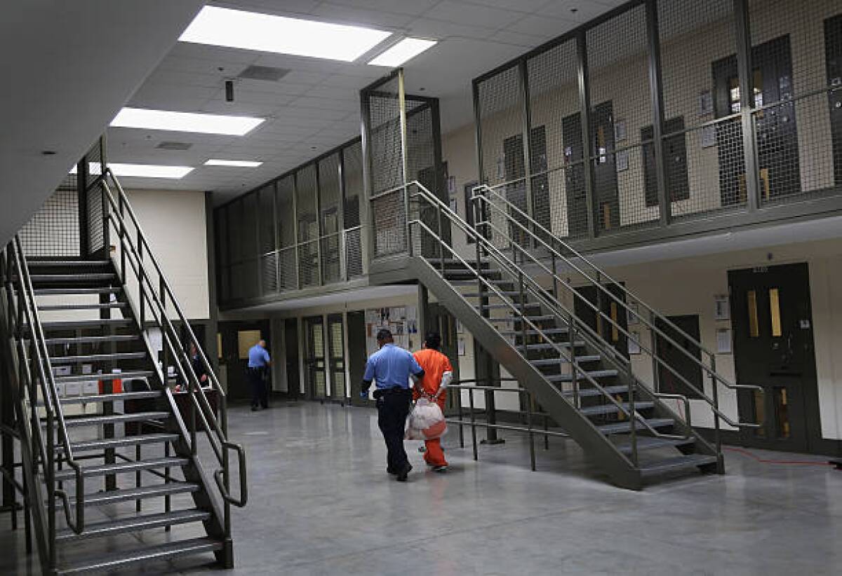 A guard in uniform, left, escorts a man dressed in orange and holding a bag near a metal staircase in a building 