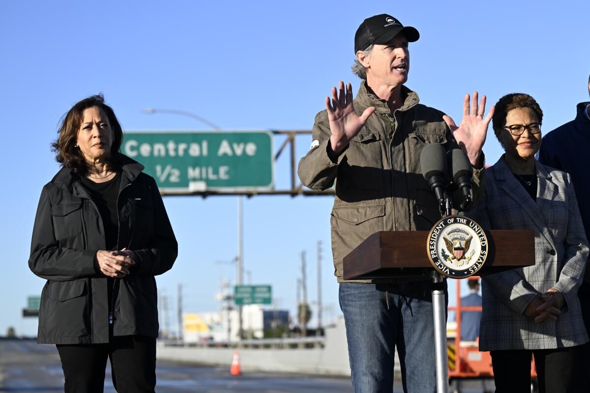 Gavin Newsom, in jacket and billed cap, and Kamala Harris and Karen Bass, in jackets, stand outdoors near a freeway exit.