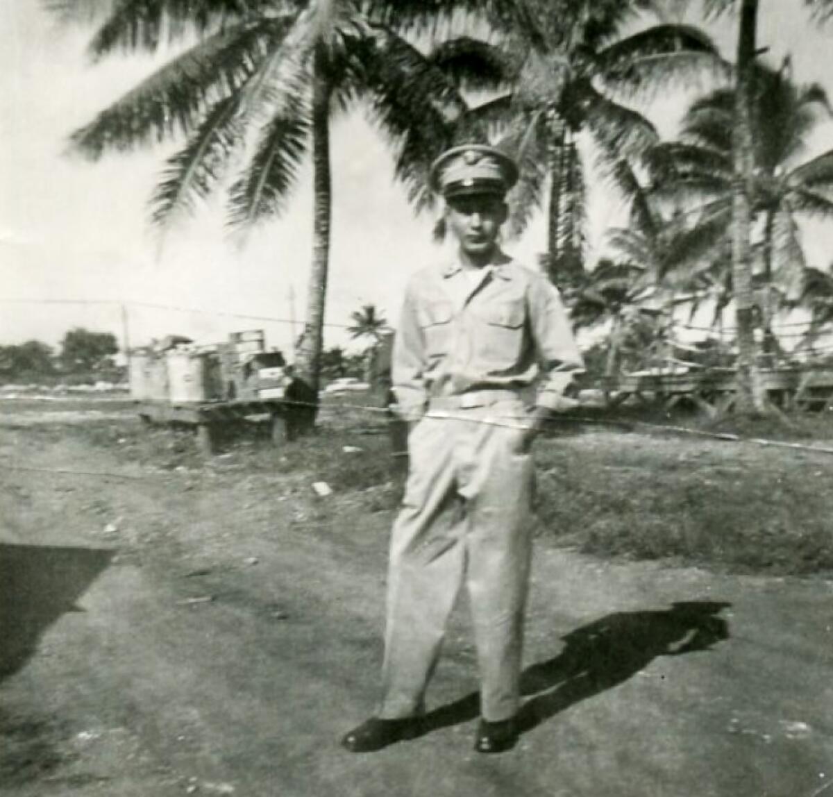 A black-and-white image of a young man in a military uniform standing in front of palm trees.