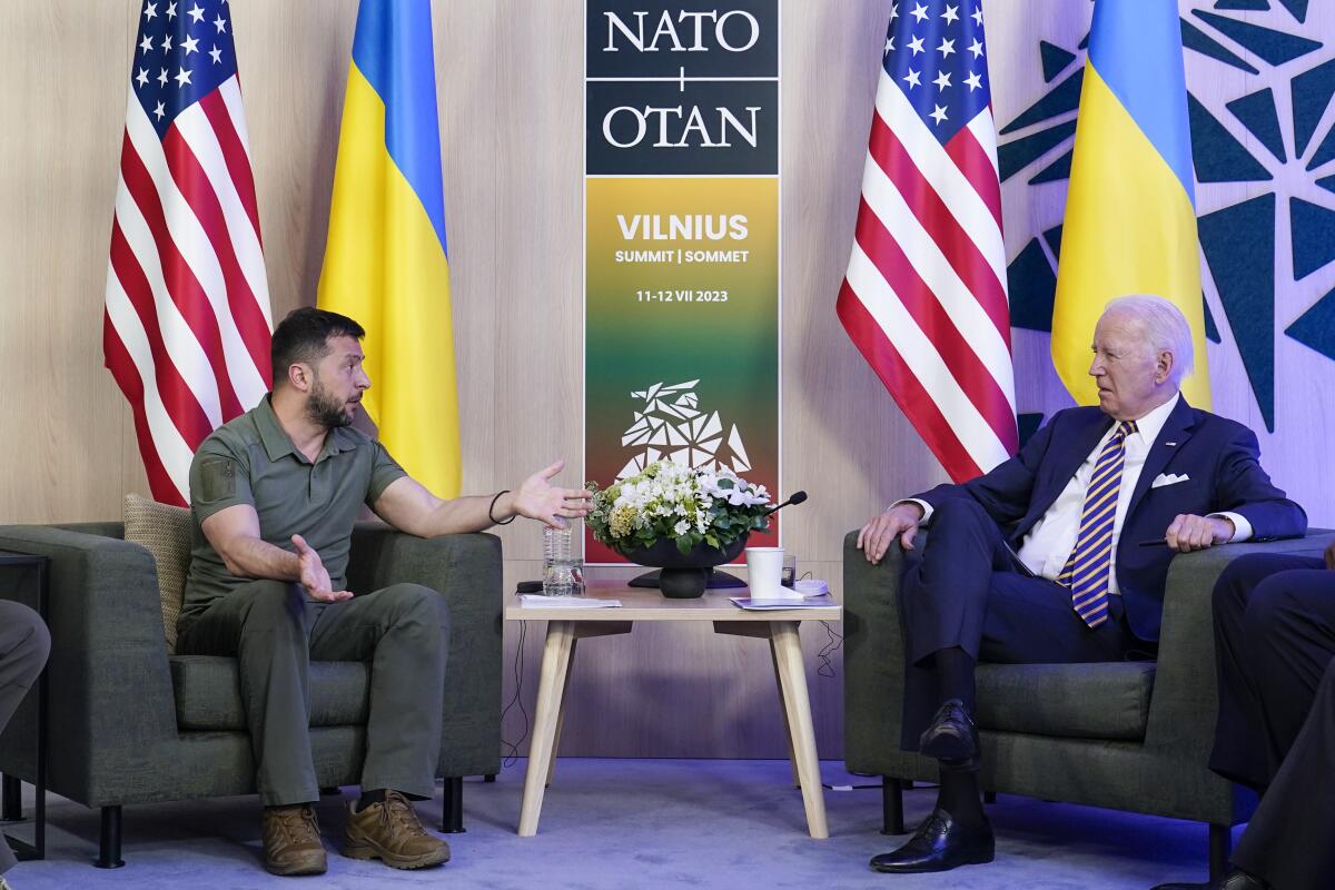 President Biden sits in a chair across from Ukraine's President Volodymyr Zelensky with flags behind them.