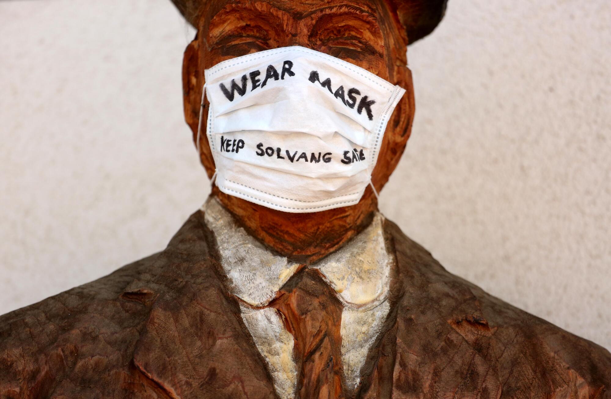 A wooden sculpture of one of the founders of Solvang wears a surgical mask with the message, "Wear Mask - Keep Solvang Safe," in front of the Copenhagen House in the California town.