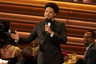 A man in a tuxedo holds a microphone among people in a ballroom