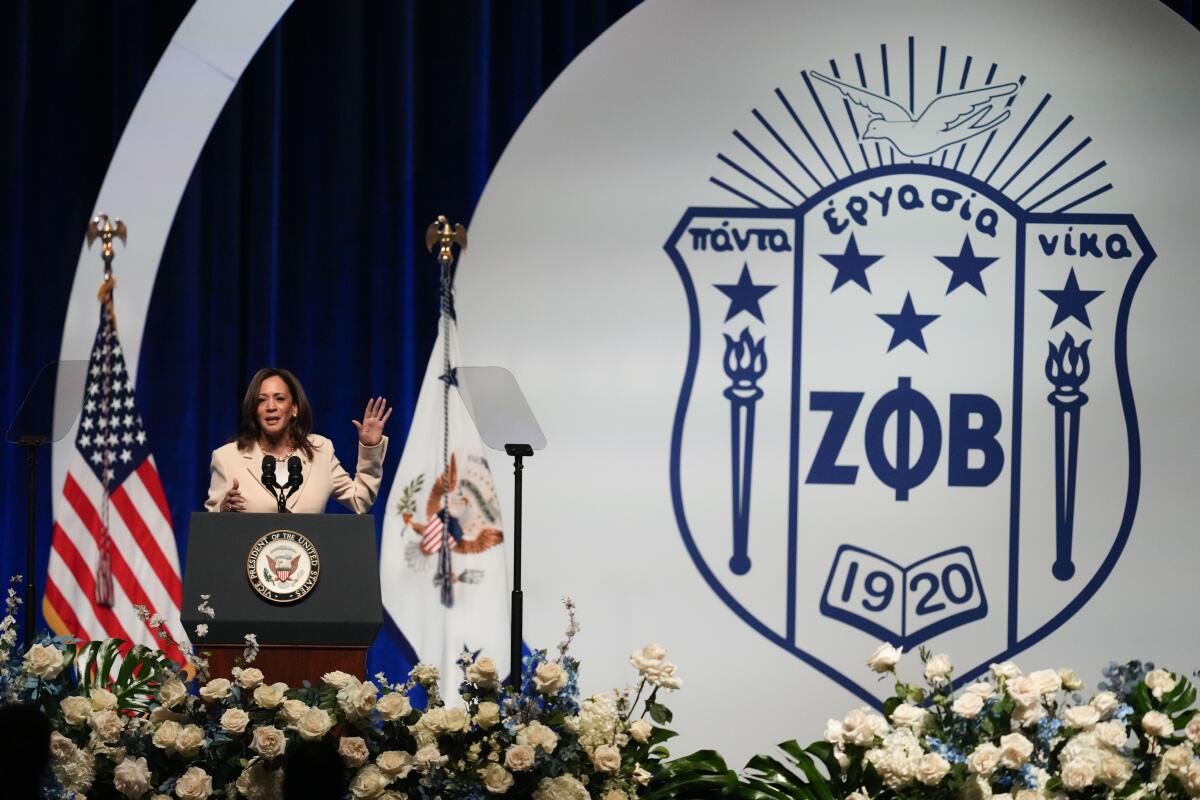 Vice President Kamala Harris, in a white suit, speaks on a stage decorated with white flowers and a Zeta Phi Beta insignia