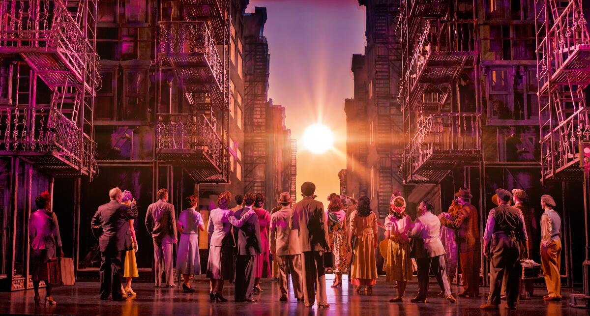 Several people in suits, dresses and work clothes face the sun between buildings in "New York, New York" on stage.