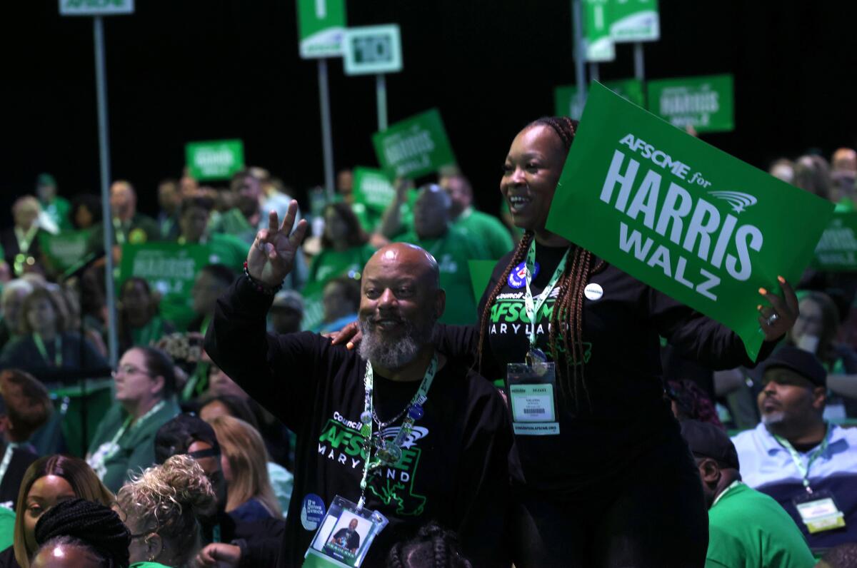 2 people, one with a green "AFSCME for Harris Walz" sign, stand in the foreground of a crowd, many holding the same sign