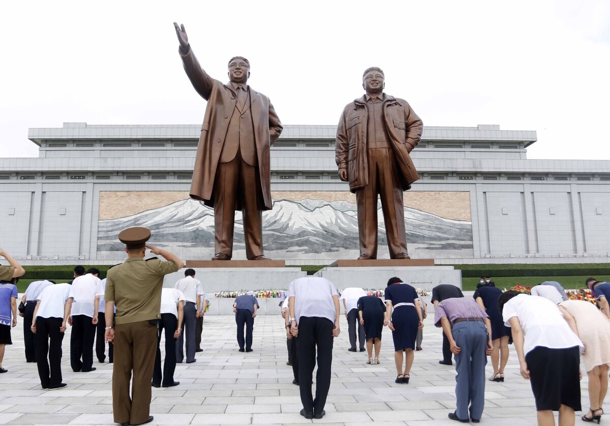 Socially distanced people bow while standing before huge bronze statues of two men

