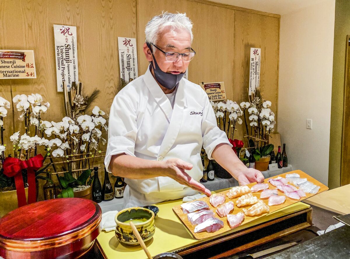 A chef stands behind a sushi counter and gestures at the raw fish before him.