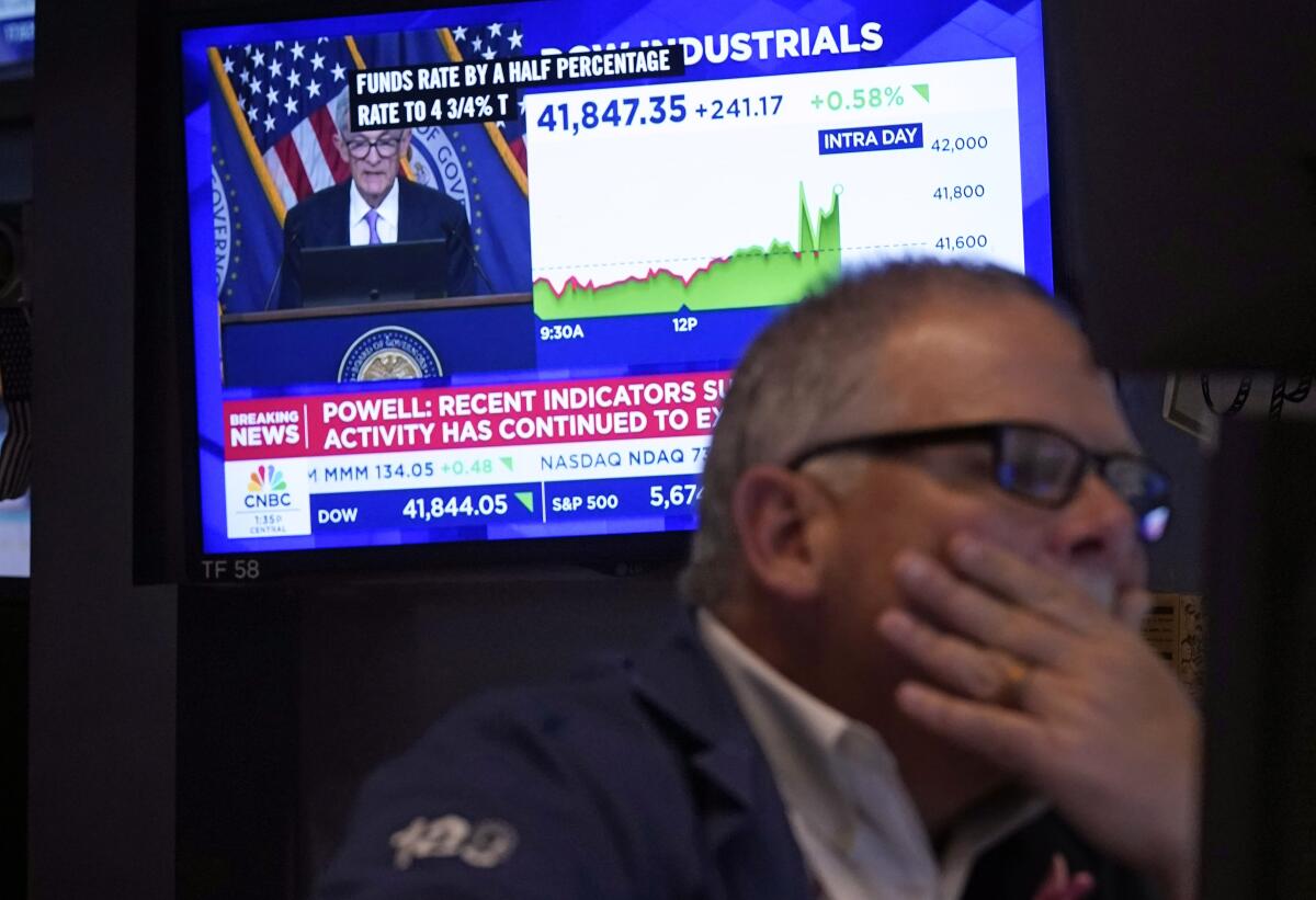 A man with glasses holds a hand over his mouth as a TV screen behind him shows stock market figures