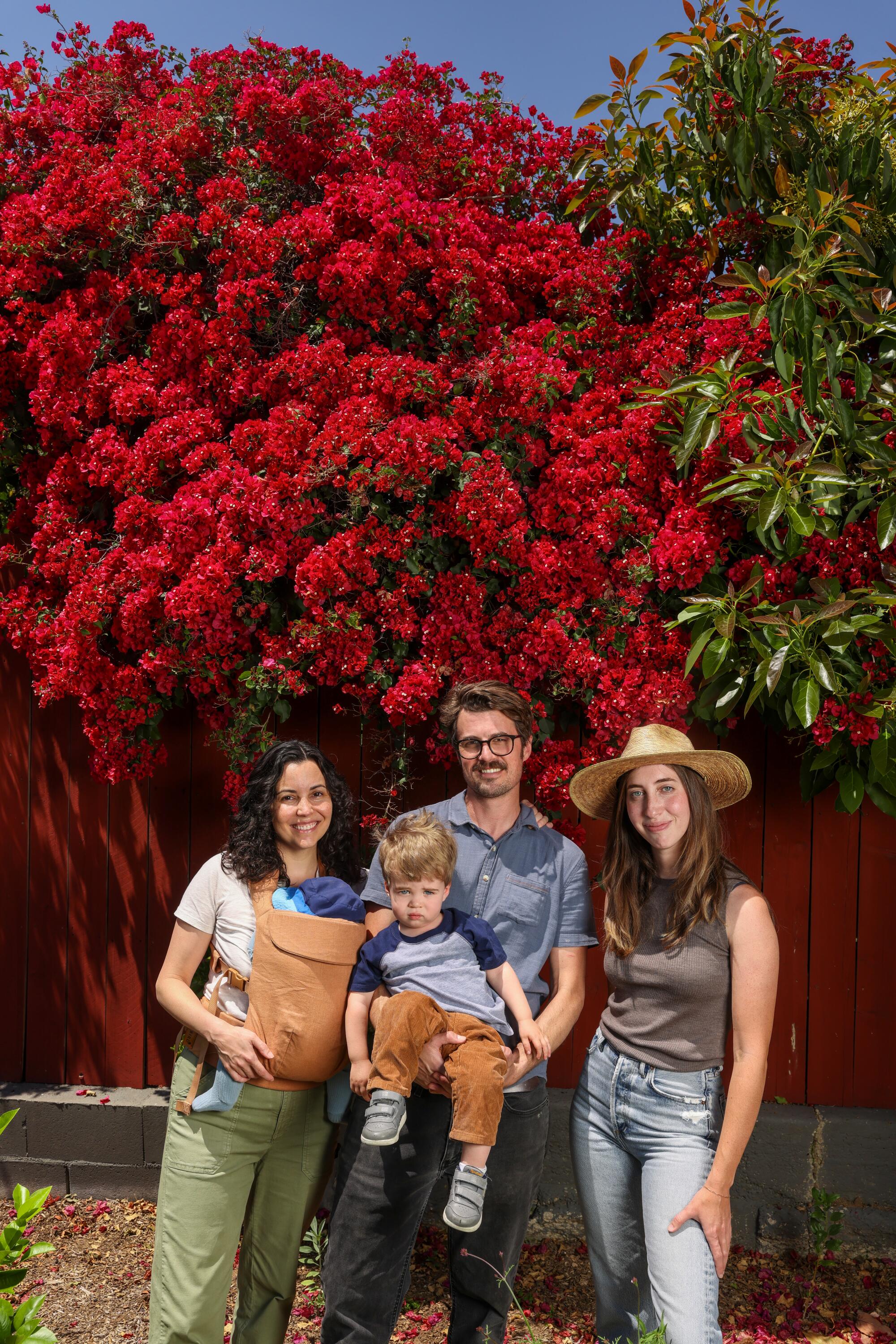 Two women, two children and one man stand for a portrait in front of a big red flowering plant.